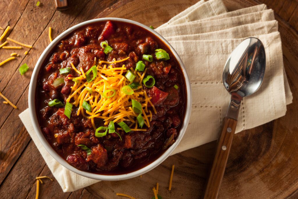 Bowl of chili topped with shredded cheddar cheese and chopped green onions, served with a spoon and napkin on a wooden table.